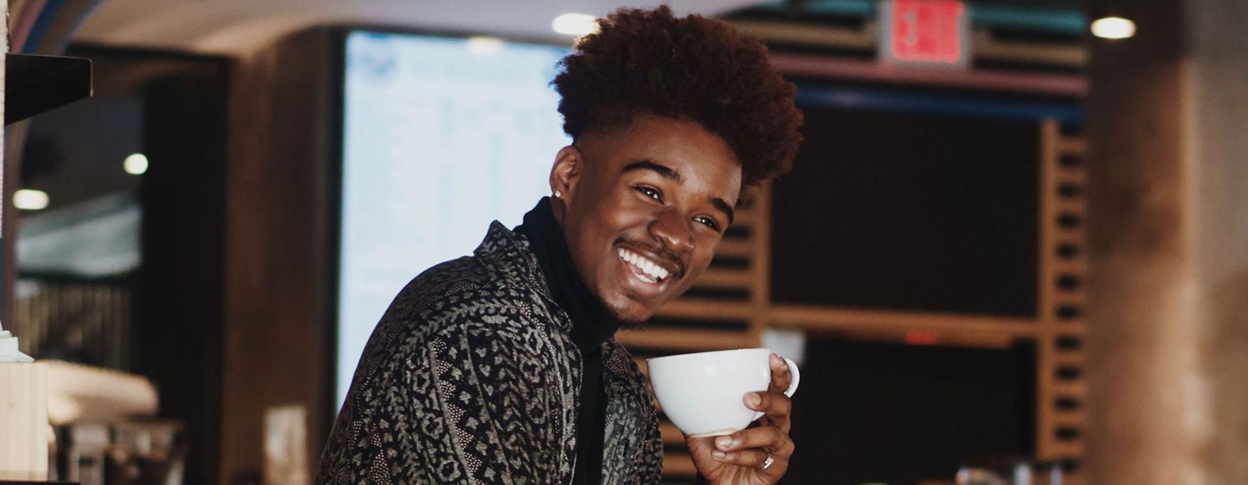 young man smiles while holding a cup of coffee in coffee shop